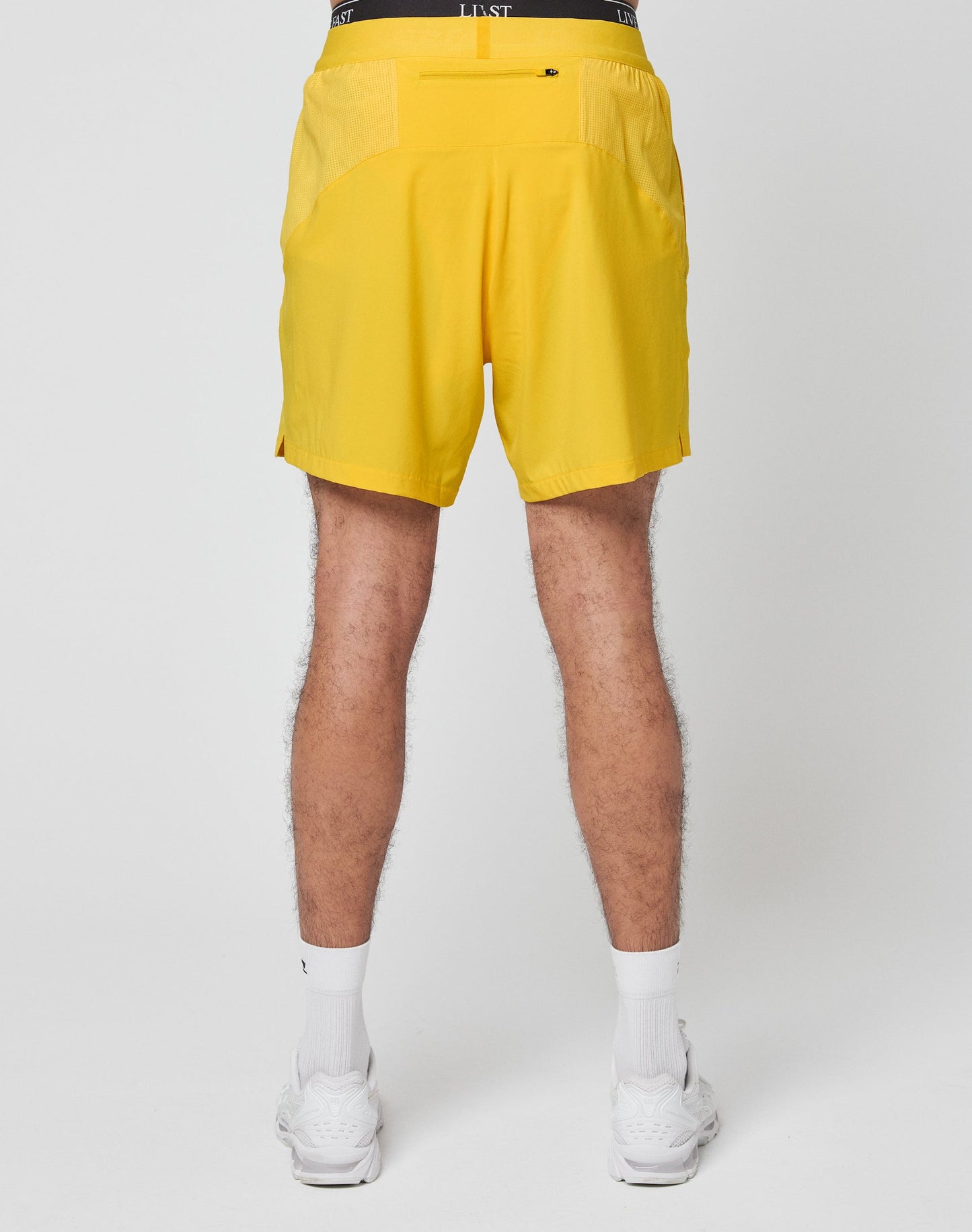 Someone stands against a white background wearing LIVE FAST DIE YOUNG’s bright yellow Live Fast Performance Shorts with a black waistband that reads LIVE FAST, paired with white socks and white sneakers.