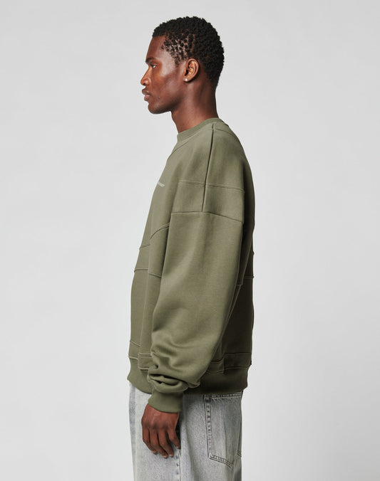 A man in profile stands against a plain background, wearing the LFDY Wavy Sweater from LIVE FAST DIE YOUNG in olive green and light gray jeans.