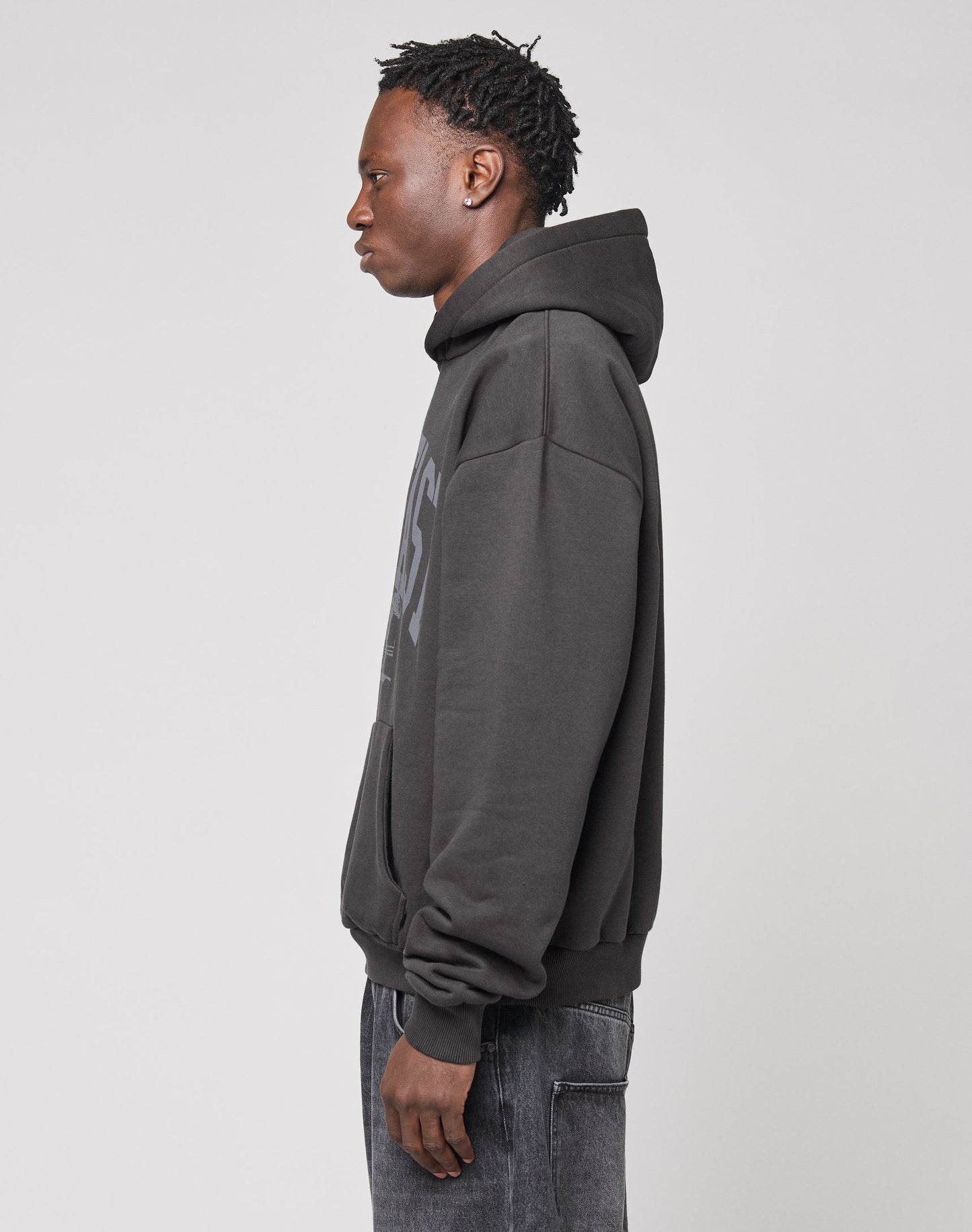 A person with short, textured hair stands in profile against a plain background, wearing the LIVE FAST DIE YOUNG Live Fast Stallion Hooded—a dark, oversized hoodie crafted from soft Baumwolle with an eye-catching front print—and dark jeans.