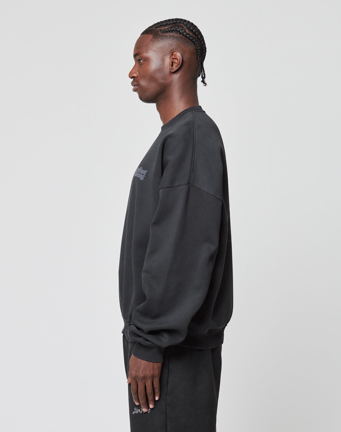 A person with braided hair stands in profile against a plain background, wearing a LIVE FAST DIE YOUNG Basic Acid Sweater in oversize fit with long sleeves, paired with matching black pants.