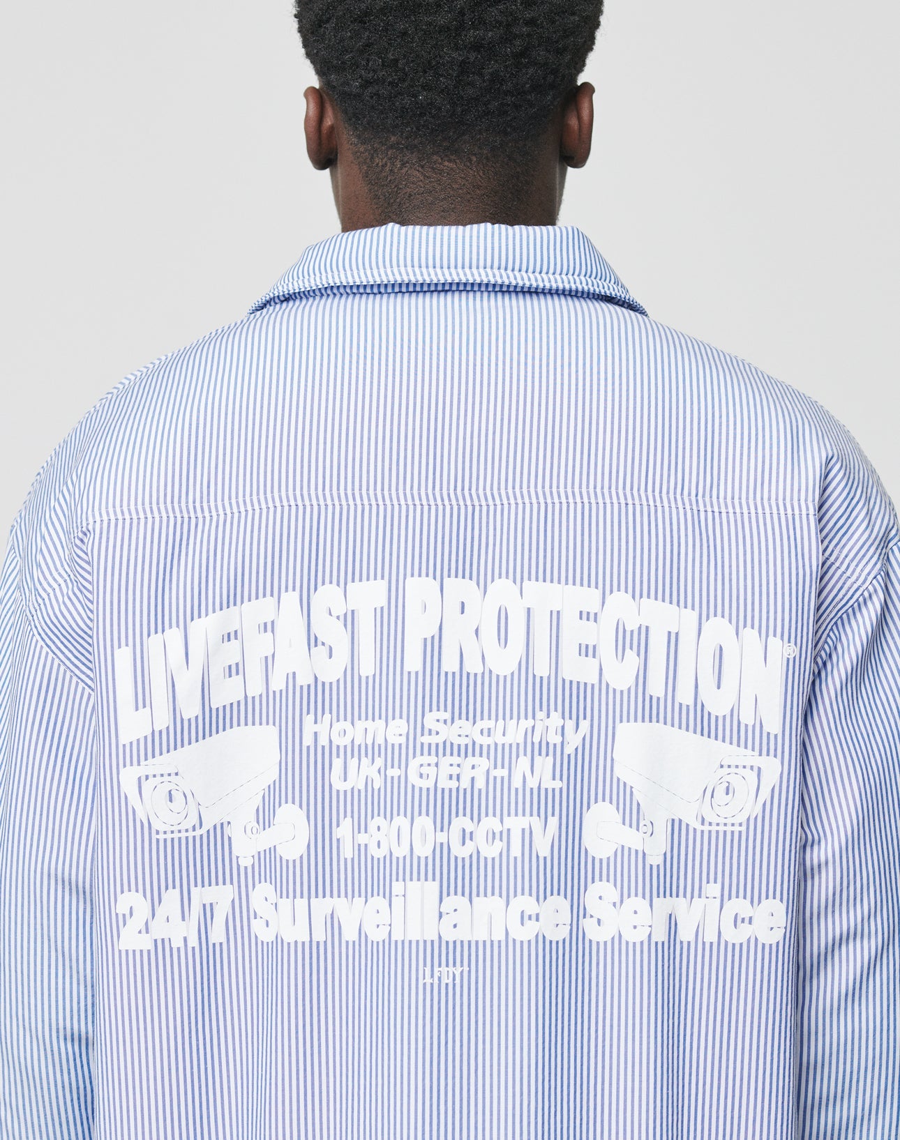 Someone wears the Padded Protection Shirt by LIVE FAST DIE YOUNG—an oversized, padded shirt with a bold backprint: LIVE FAST, PROTECTION, security cameras, 24/7 Surveillance Service, and a small Backprint Flag-Label for extra detail.