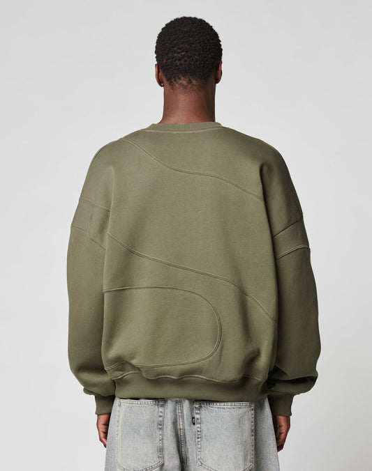 A person with short, textured hair is seen from behind in an LFDY Wavy Sweater by LIVE FAST DIE YOUNG—an oversize olive green top in soft cotton-polyester—paired with light-wash denim jeans against a plain light gray background.