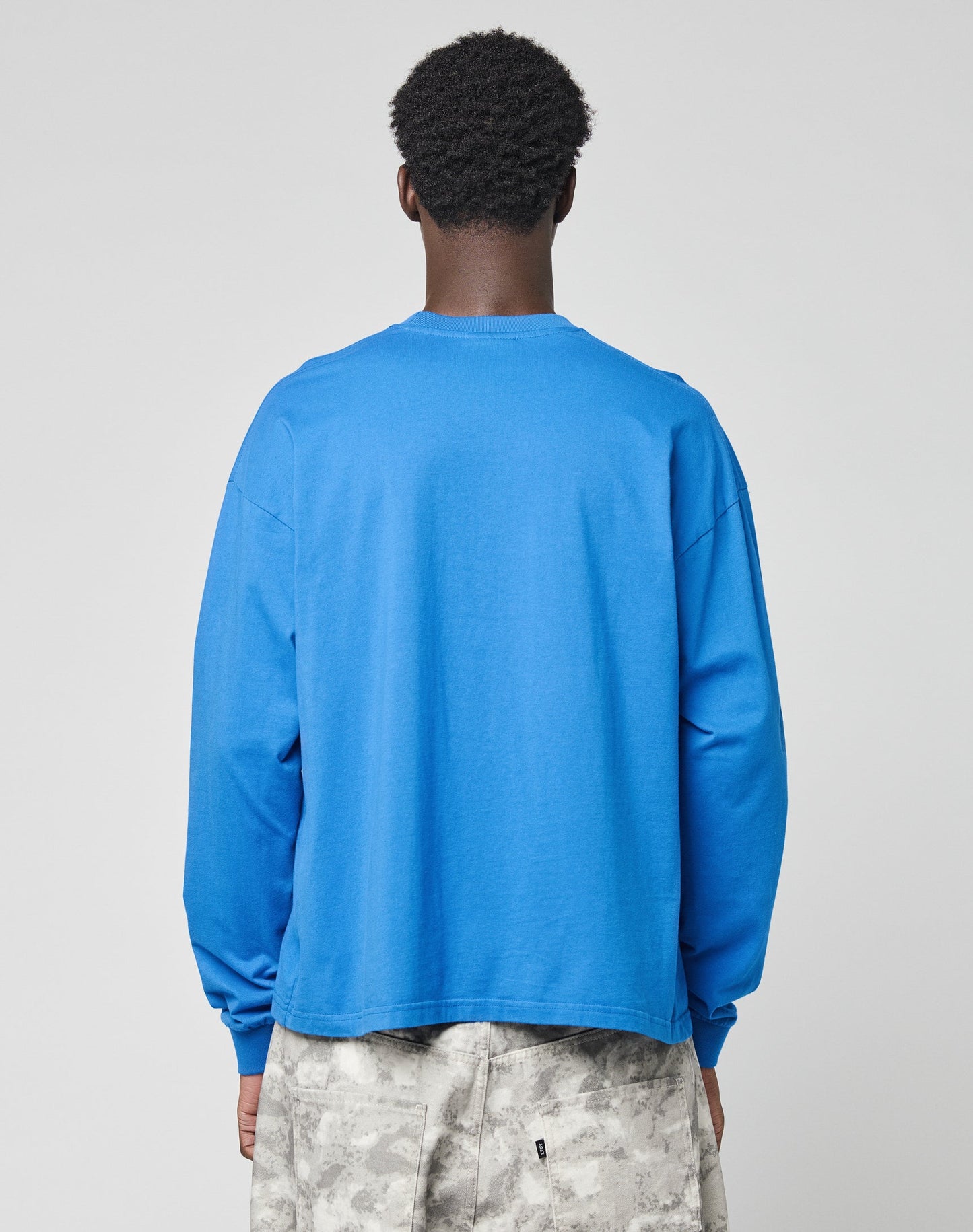 A person with short curly hair is seen from behind wearing an oversize bright blue LIVE FAST DIE YOUNG Live Fast Archive Longsleeve and light gray camouflage shorts against a plain light background.