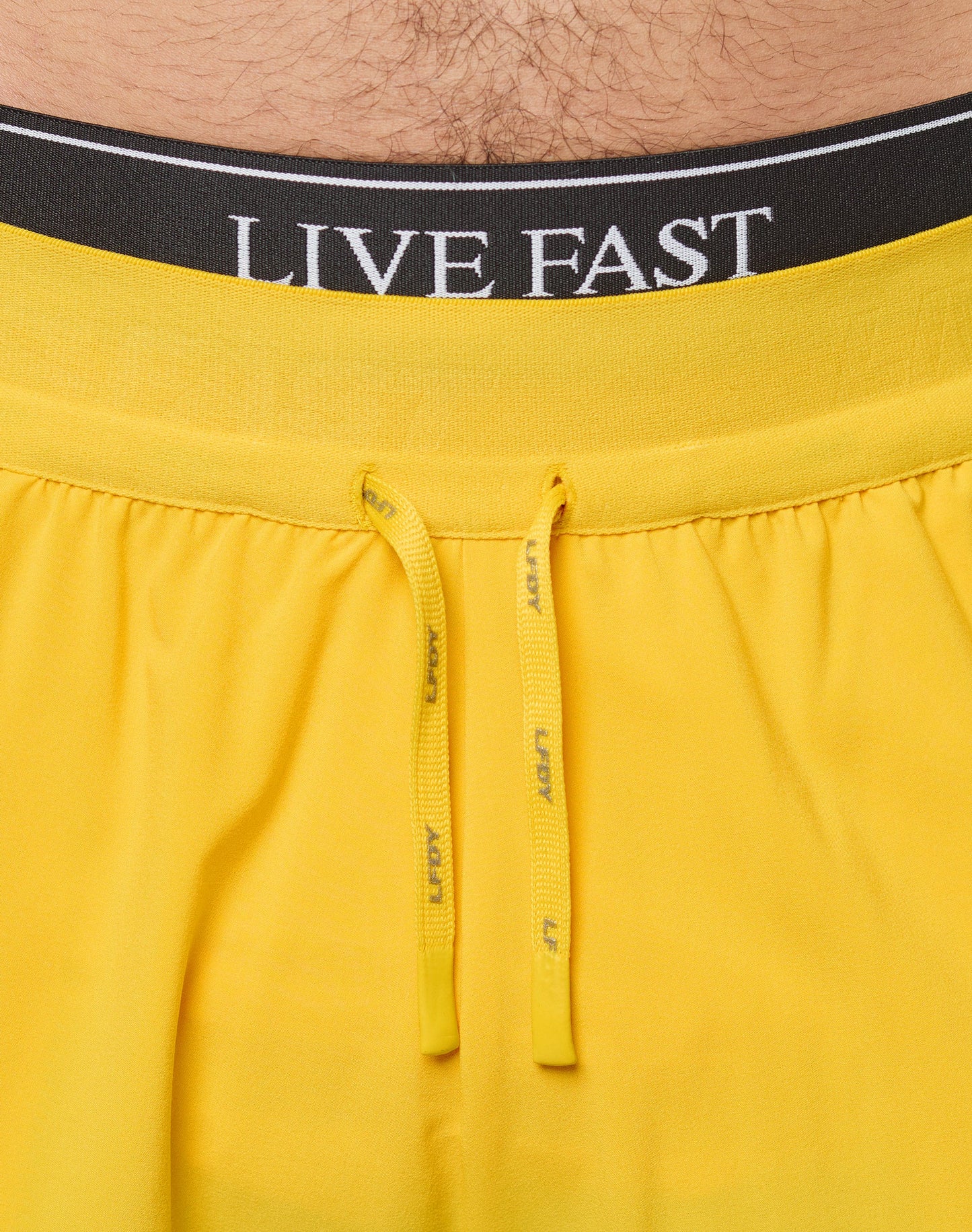 A person models the LIVE FAST DIE YOUNG Live Fast Performance Shorts in bright yellow with an elastic waistband and small reflective print near the hem, paired with white socks and athletic sneakers. The upper body is not shown.| model