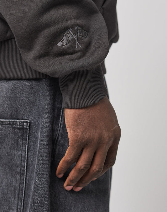 A person’s right hand hangs by their side, wearing the LIVE FAST DIE YOUNG Live Fast Stallion Hooded, a dark oversized long-sleeve top with embroidered cuffs, paired with gray denim pants against a plain light background.
