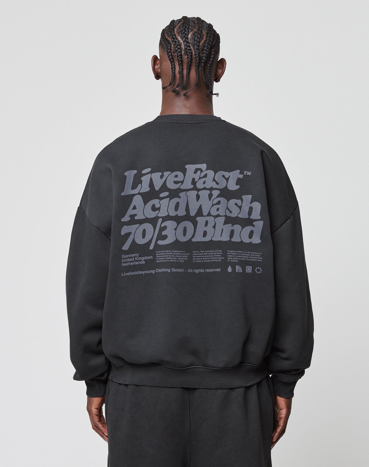 A person with braided hair is seen from behind wearing the Basic Acid Sweater by LIVE FAST DIE YOUNG, an oversized black sweatshirt with bold gray text and vintage-wash details, crafted from a soft cotton-polyester blend.