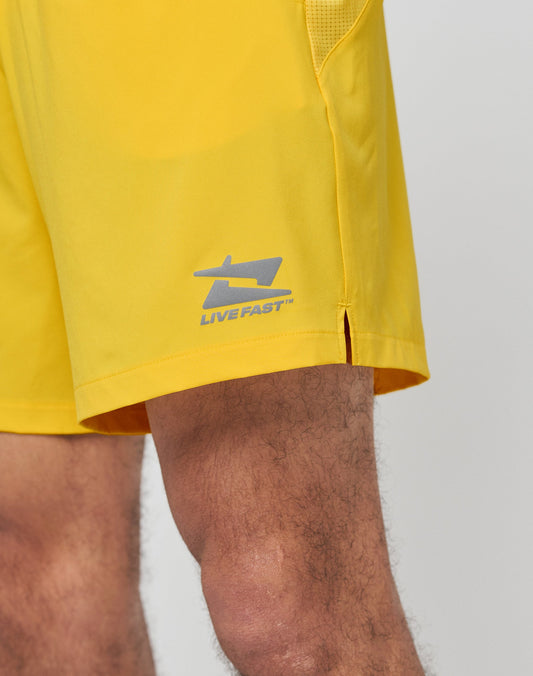 A close-up shows only the lower leg of a person wearing bright yellow Live Fast Performance Shorts by LIVE FAST DIE YOUNG, featuring a Regular Fit, elastic waistband, small slit, and silver reflective LIVEFAST logo on the side.