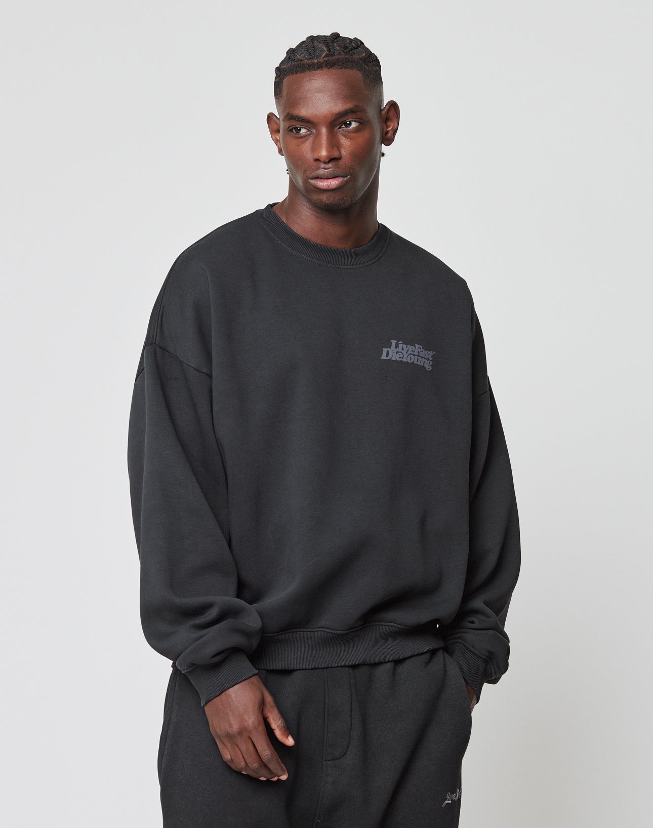 A man stands against a plain light background, wearing the LIVE FAST DIE YOUNG Basic Acid Sweater and black pants. He gazes slightly to the side with a neutral expression, one hand by his side and the other in his pocket.