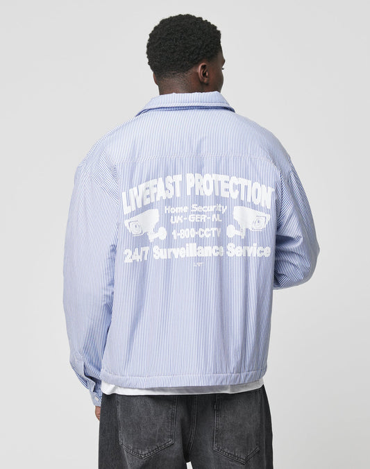 A person wearing a blue and white striped Padded Protection Shirt by LIVE FAST DIE YOUNG stands facing away. The backprint flag-label shows bold white LIVE FAST PROTECTION text with two crossed wrenches illustrated below.