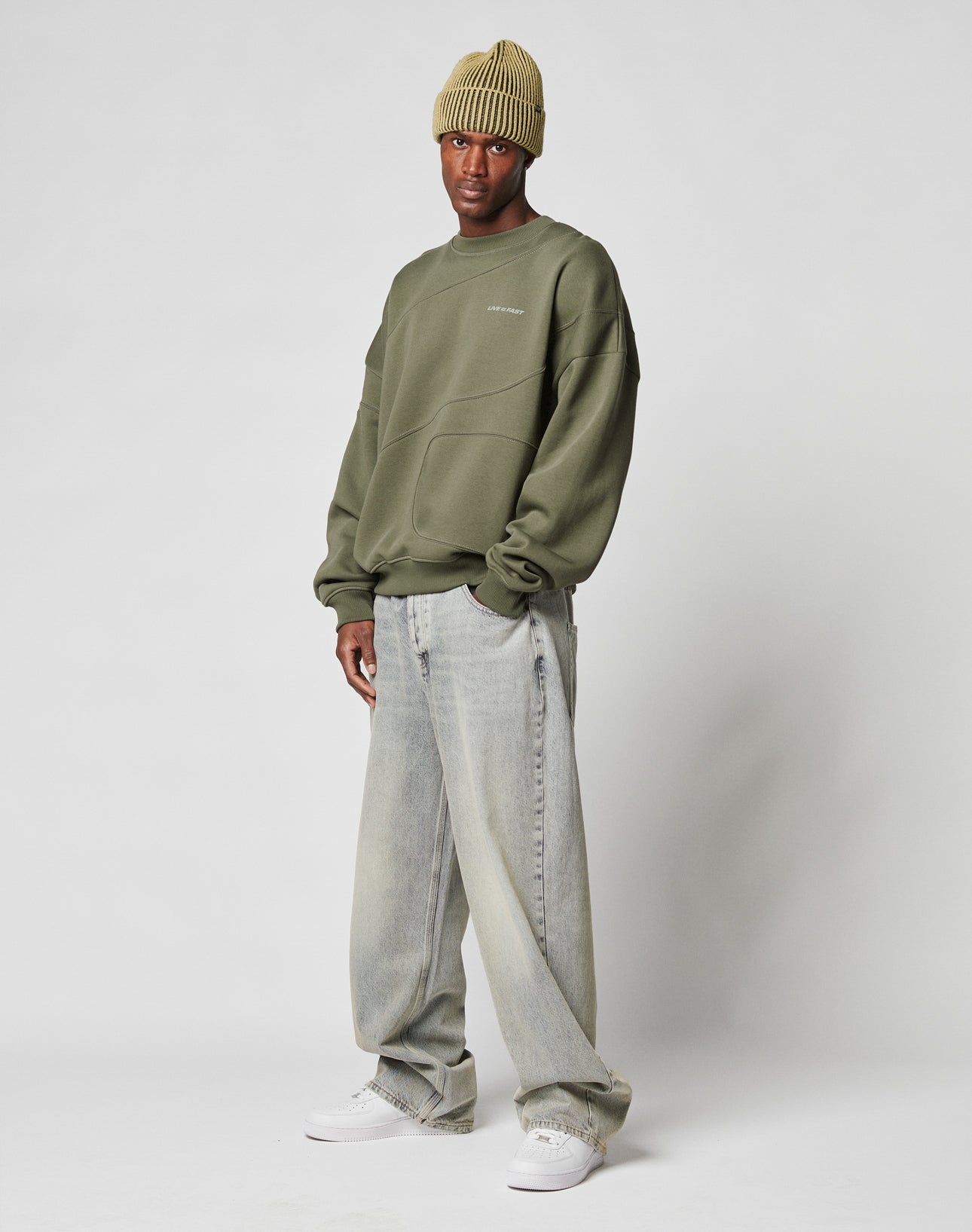 A man wearing the LIVE FAST DIE YOUNG LFDY Wavy Sweater in olive green, a light green ribbed beanie, loose faded jeans, and white sneakers stands against a plain white background.