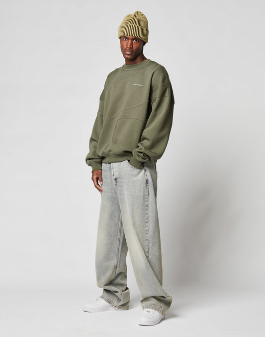 A man wearing the LIVE FAST DIE YOUNG LFDY Wavy Sweater in olive green, a light green ribbed beanie, loose faded jeans, and white sneakers stands against a plain white background.