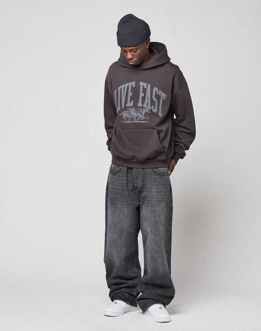 Wearing a dark beanie, the person stands against a light background in loose dark jeans, white sneakers, and an Oversize Fit LIVE FAST DIE YOUNG Live Fast Stallion Hooded hoodie with hands in their pockets, looking down.