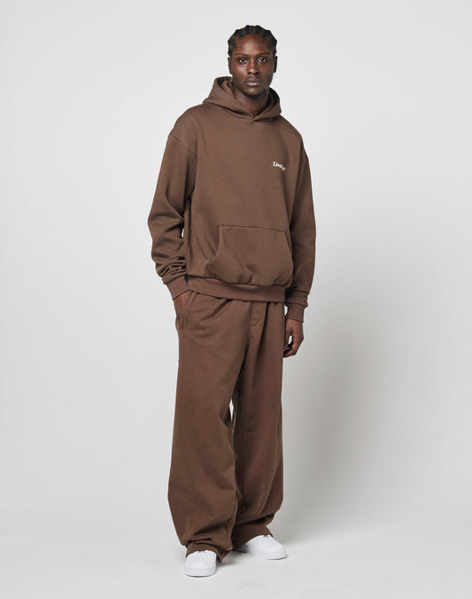A person with a neutral expression stands against a plain background wearing the LIVE FAST DIE YOUNG Basic 360 Hooded brown oversize hoodie and matching wide-leg polyester pants, paired with white sneakers and hands by their sides.