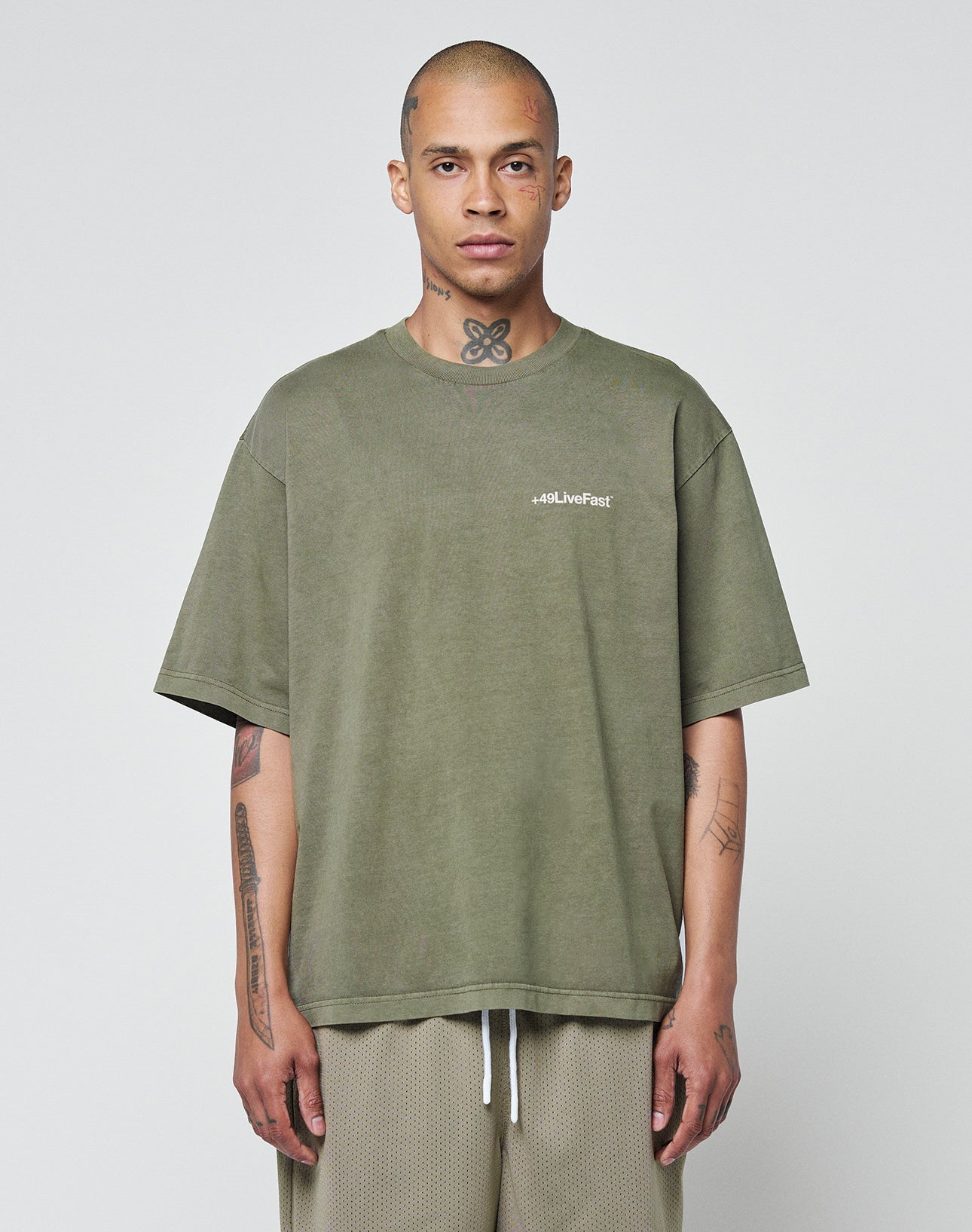A tattooed person stands against a plain background, wearing the LIVE FAST DIE YOUNG Basic +49 Chest Tee—an olive-green, 100% Baumwolle oversize t-shirt with a #QLLiveFast logo on the left chest—and light-colored mesh shorts.