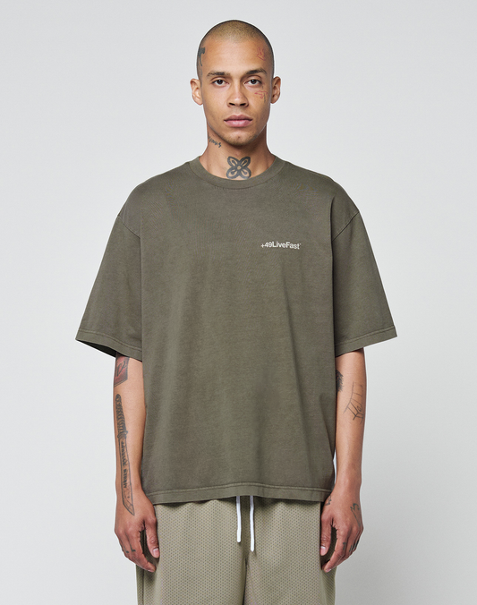 A person with a shaved head and tattoos stands facing forward, wearing the LIVE FAST DIE YOUNG Basic +49 Chest Tee in olive green with logo print and light mesh shorts, set against a plain light background. | model