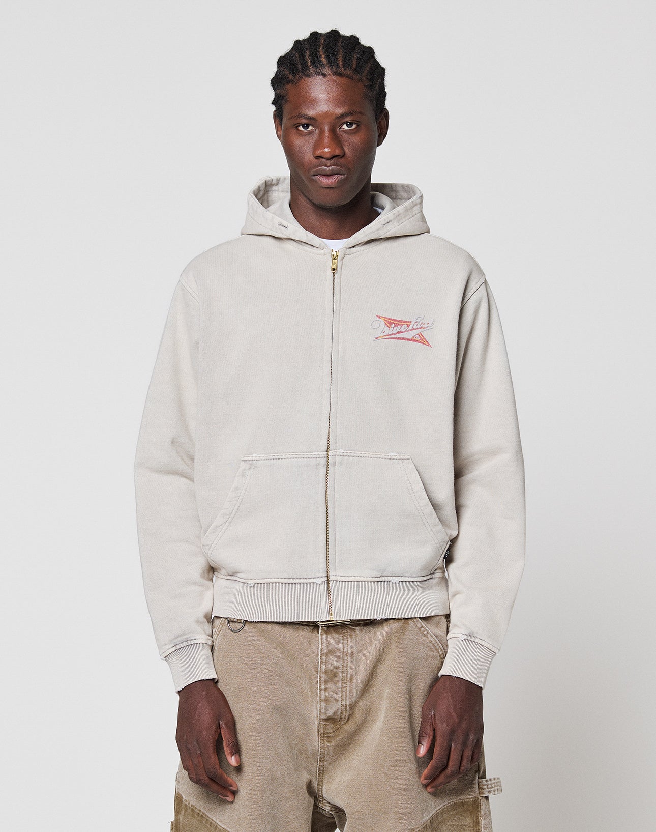 A man with braided hair wears a light gray LIVE FAST DIE YOUNG LFDY Corp. Hooded Zip with a small red and white chest graphic and tan cargo pants, standing against a plain white background. | model