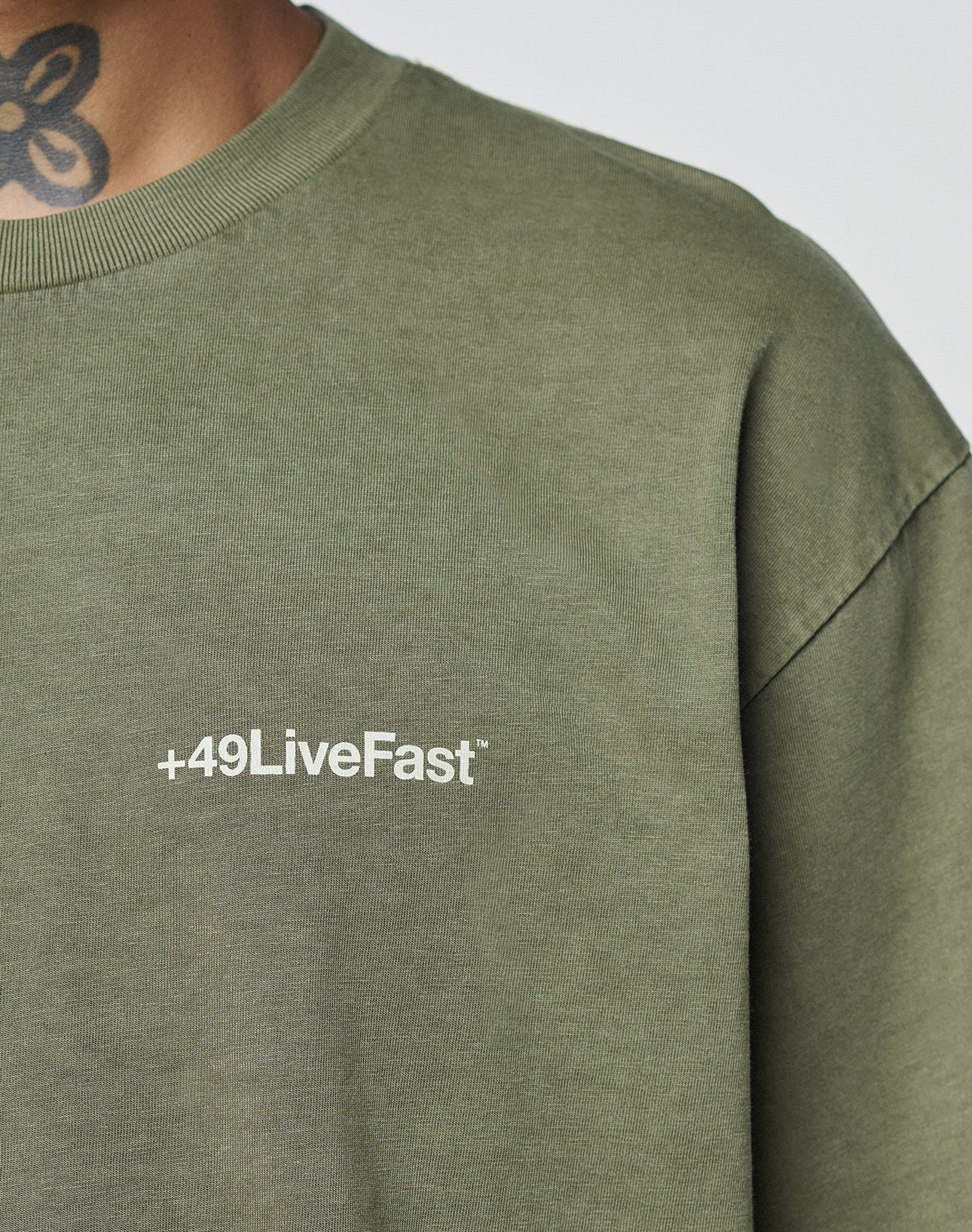 Someone wearing the olive green LIVE FAST DIE YOUNG Basic +49 Chest Tee, featuring a white +49LiveFast logo on the chest. The 100% Baumwolle tee has an oversize fit, with part of a neck tattoo visible at the top of the image.