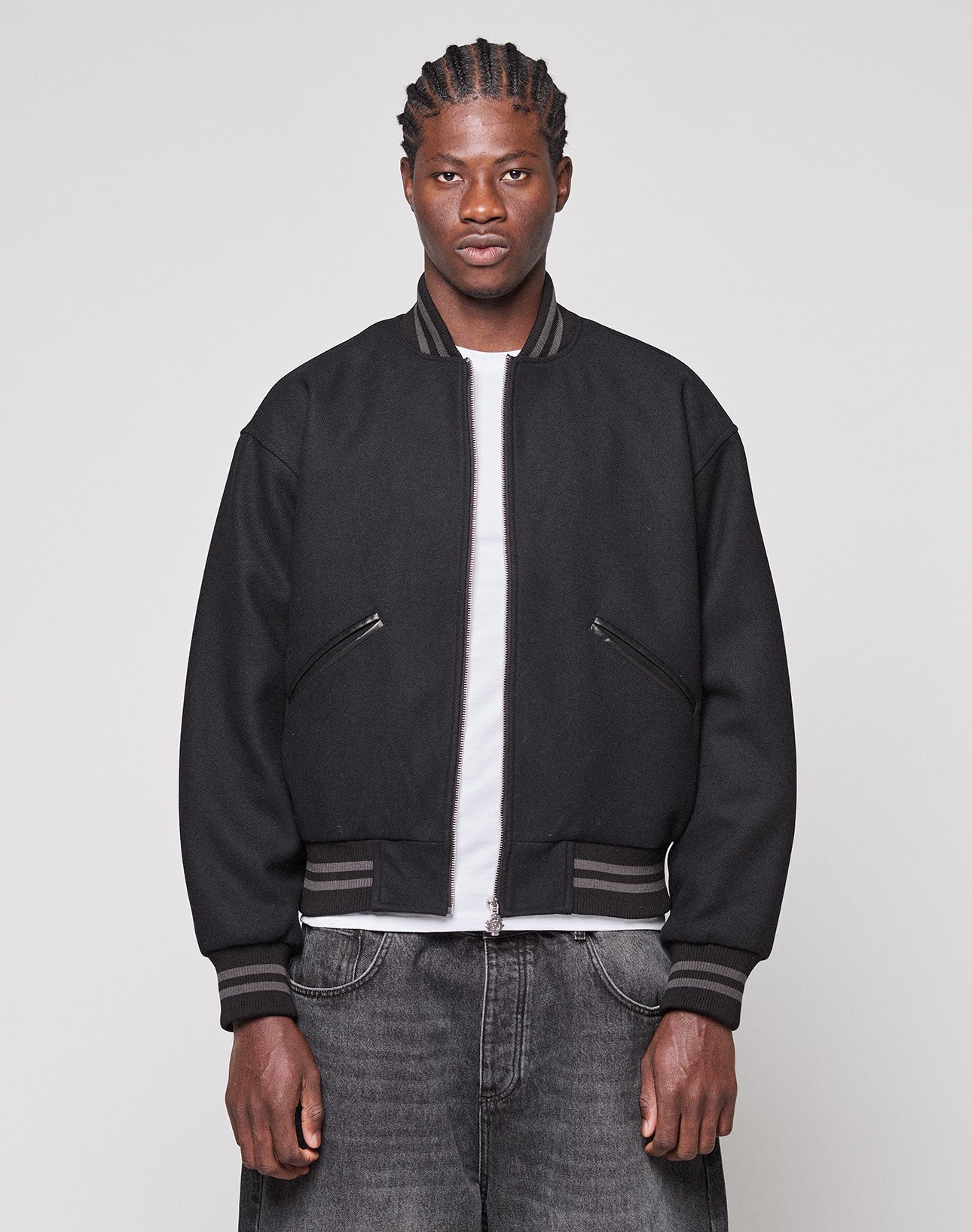 A person with braided hair wears the LIVE FAST DIE YOUNG Live Fast College Jacket, featuring a metal zipper, over a white T-shirt and dark jeans, standing against a plain light gray background and looking directly at the camera.