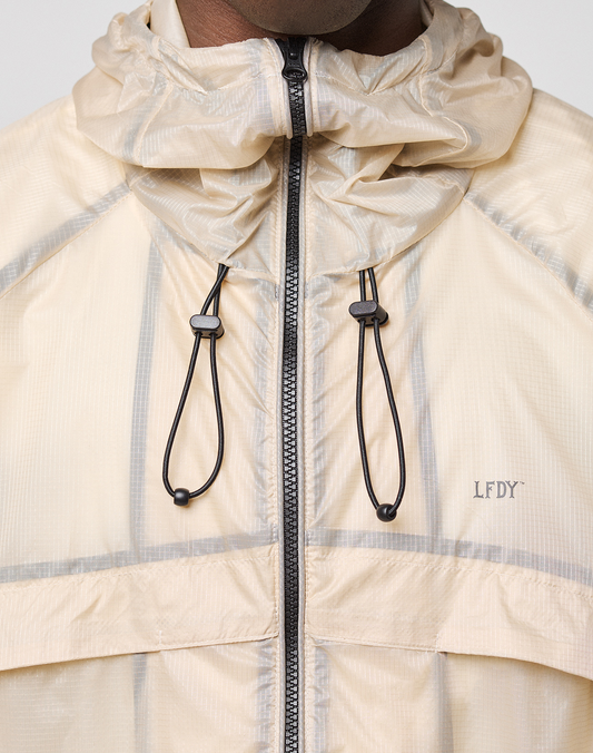 Close-up of a person wearing the beige Live Fast Nylon Jacket by LIVE FAST DIE YOUNG, featuring black drawstrings, a zipper, LFDY logo on the left chest, and a transparent outer layer. Only the lower face and neck are visible.