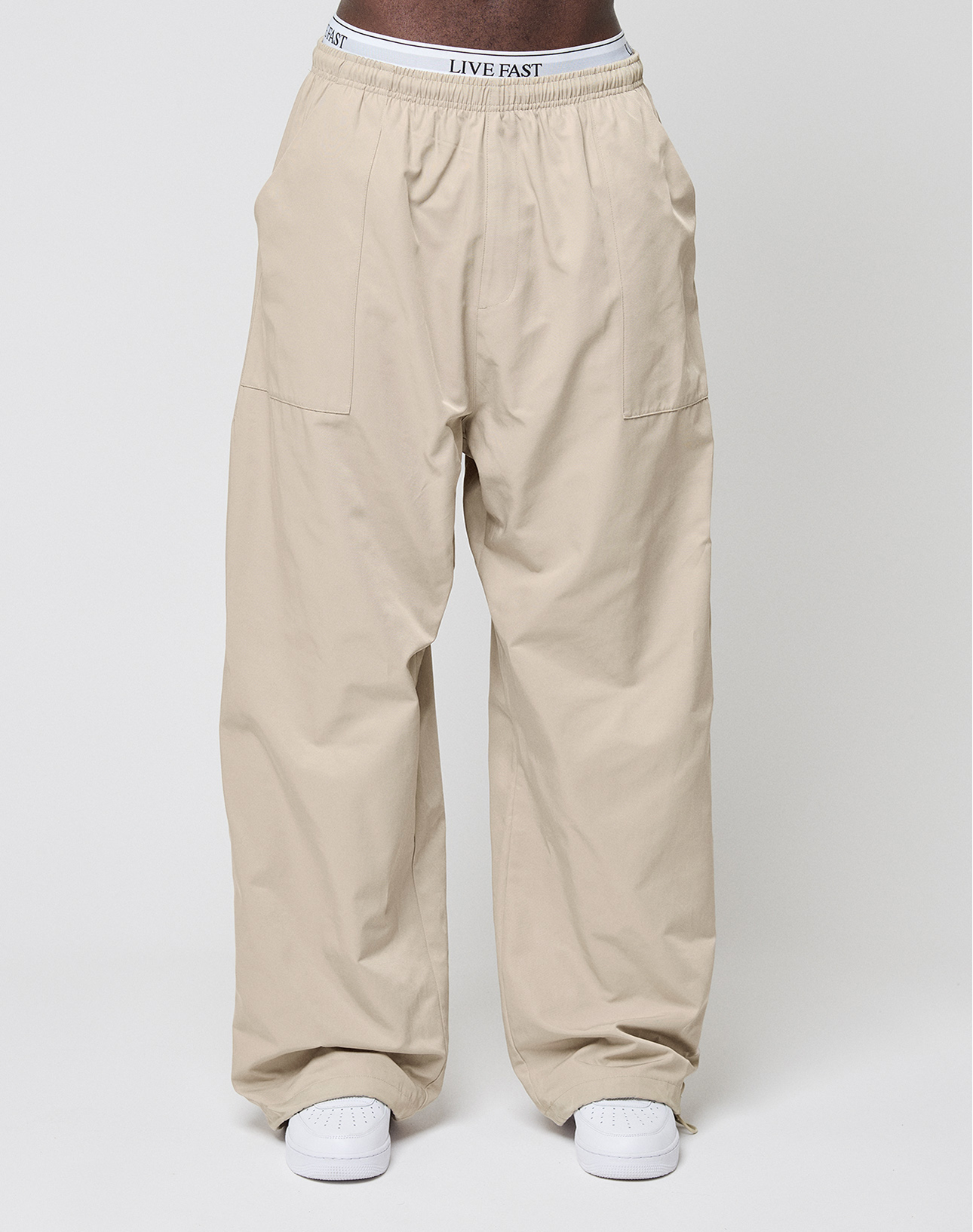 A person wears LIVE FAST DIE YOUNG Baggy Nylon Pants with aufgesetzte Taschen, white sneakers, and a white elastic waistband that reads LIVE FAST against a plain light background.