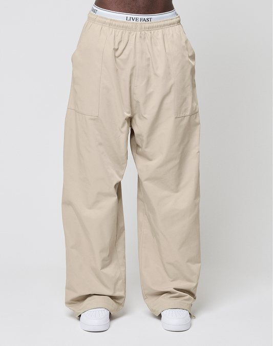 A person wears LIVE FAST DIE YOUNG Baggy Nylon Pants with aufgesetzte Taschen, white sneakers, and a white elastic waistband that reads LIVE FAST against a plain light background.