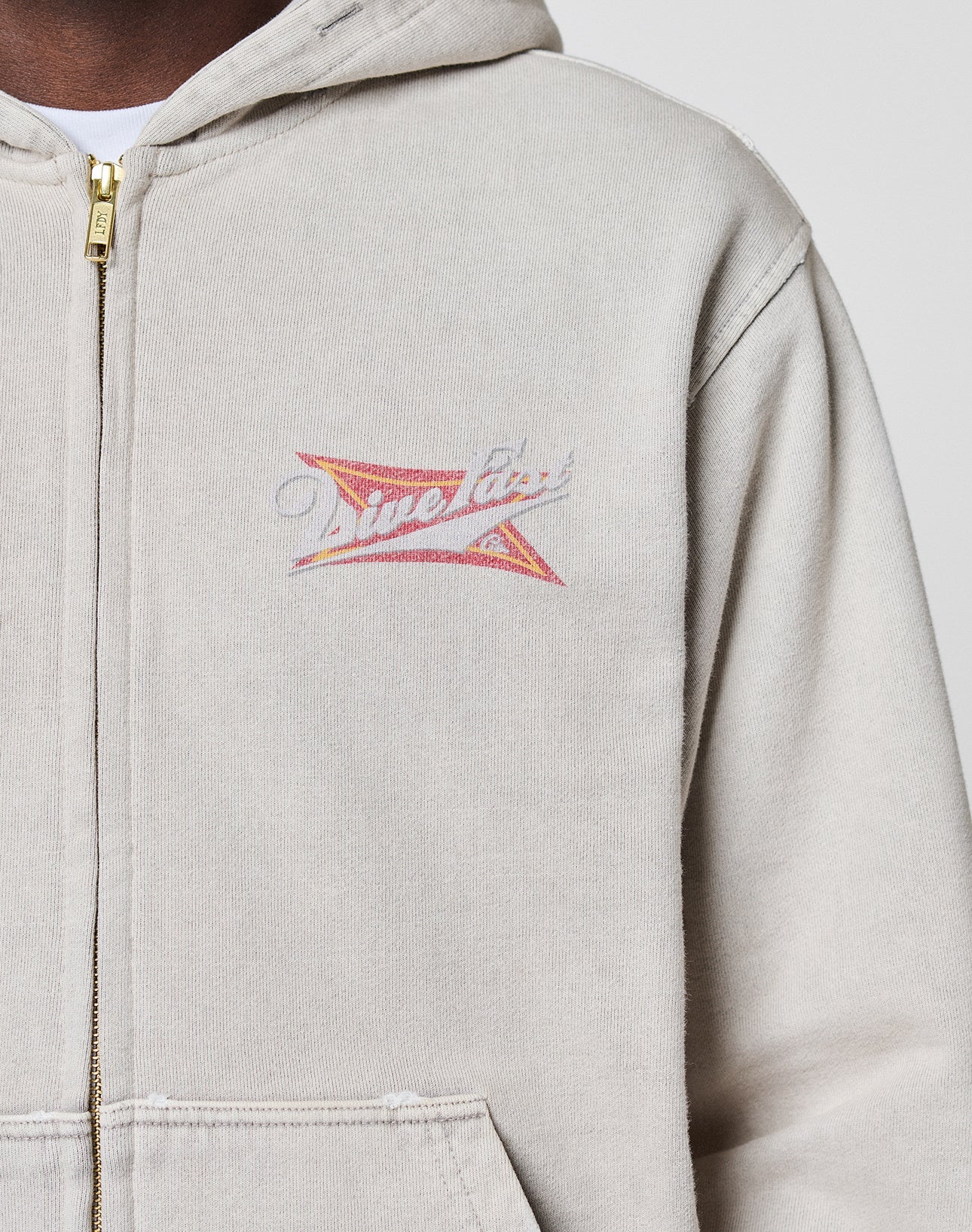 A person wearing a LIVE FAST DIE YOUNG LFDY Corp. Hooded Zip in light beige 100% Baumwolle with a gold zipper, featuring a small colorful graphic and "Time Flies" on the left chest. Only the upper torso is visible.