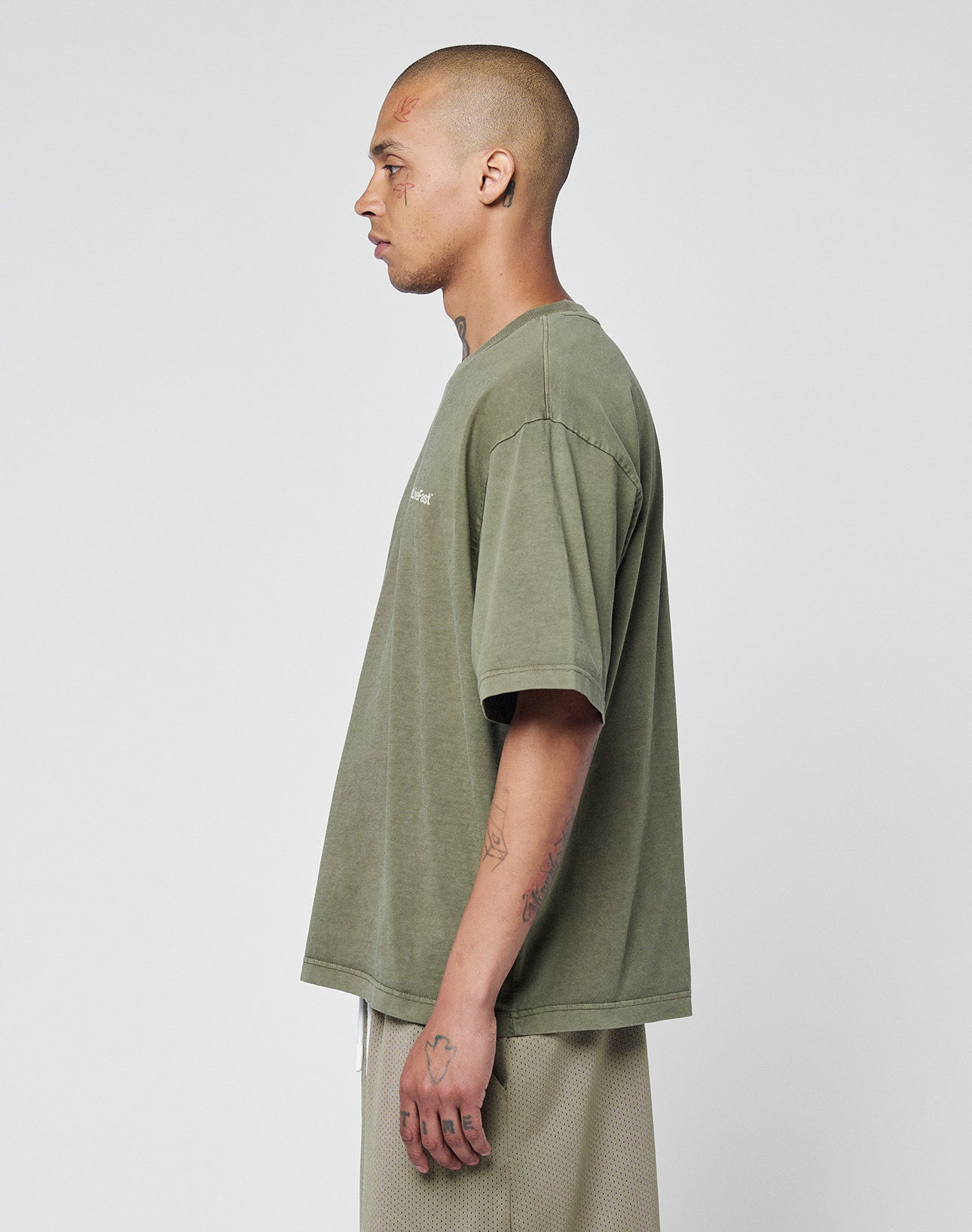 A person with a shaved head and visible tattoos wears the LIVE FAST DIE YOUNG Basic +49 Chest Tee in olive green with beige shorts, standing in profile against a plain light background.