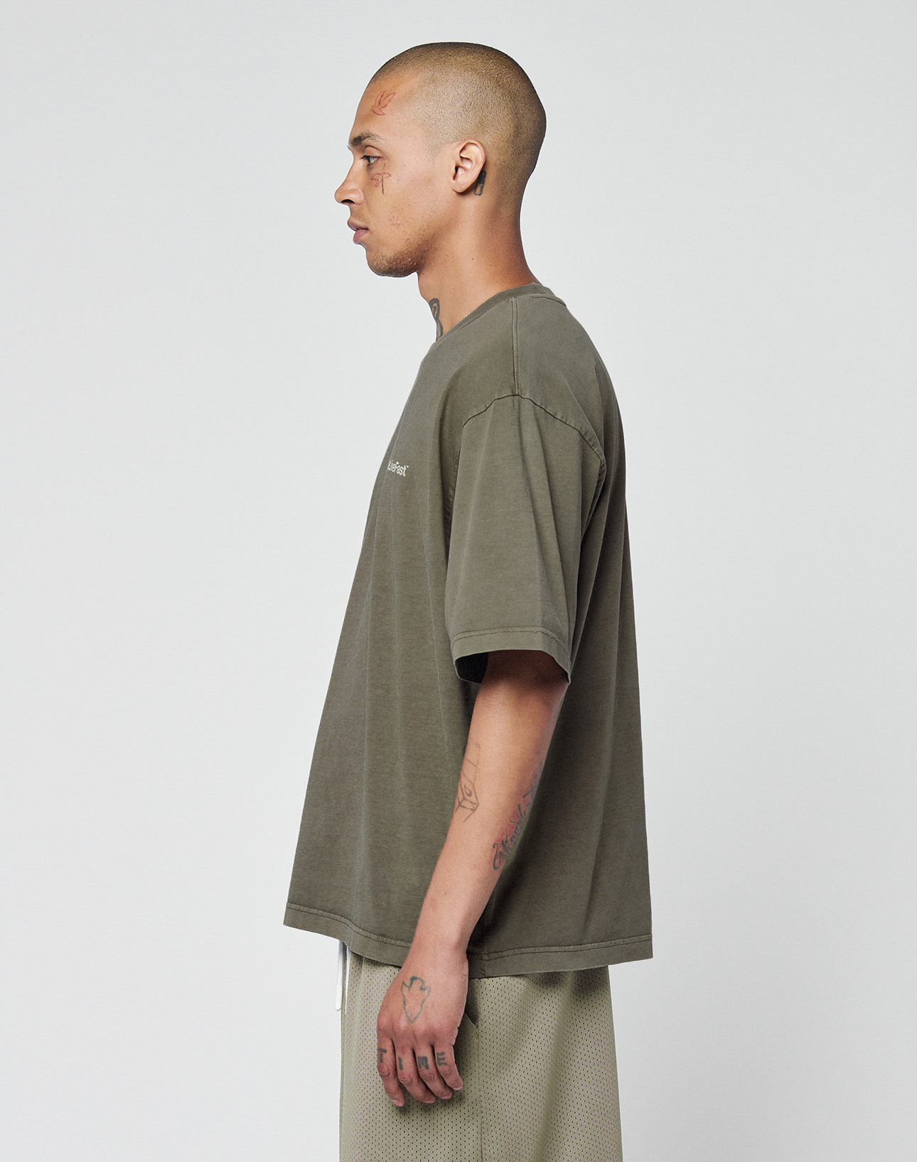 A person with a shaved head and tattoos stands in profile, wearing the LIVE FAST DIE YOUNG Basic +49 Chest Tee in olive green and light-colored shorts against a plain white background.