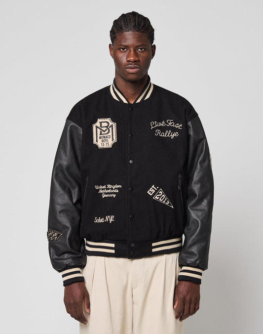 A man in cream pleated trousers wears the LFDY x Schott NYC Varsity Jacket by LIVE FAST DIE YOUNG, featuring black fabric with cream patches and writing, as he stands against a plain light gray background with a neutral expression.