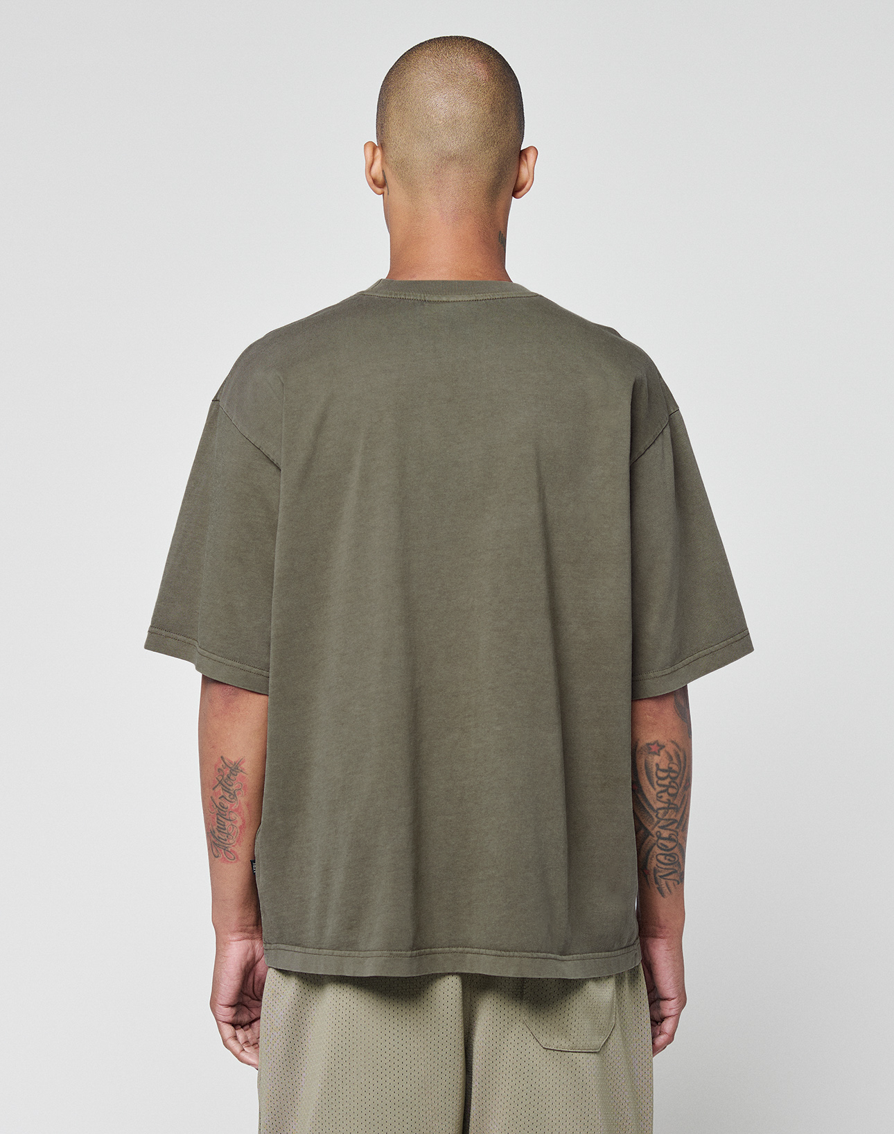 A person with a shaved head stands with their back to the camera, wearing an olive-green LIVE FAST DIE YOUNG Basic +49 Chest Tee and mesh shorts. Tattoos cover their forearms, set against a plain white background that highlights their style.