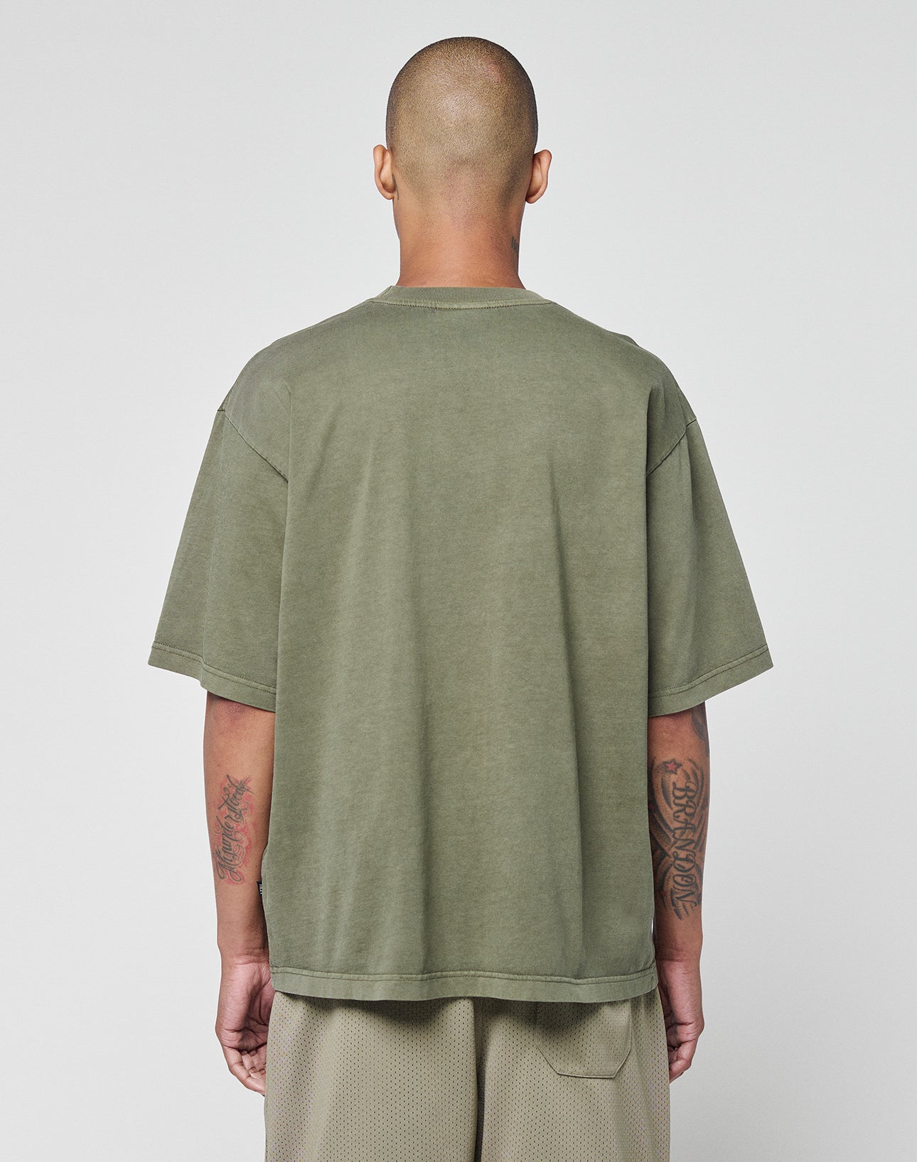 A person with a shaved head faces away, wearing a LIVE FAST DIE YOUNG Basic +49 Chest Tee in olive green and light beige shorts, revealing forearm tattoos against a plain light background.