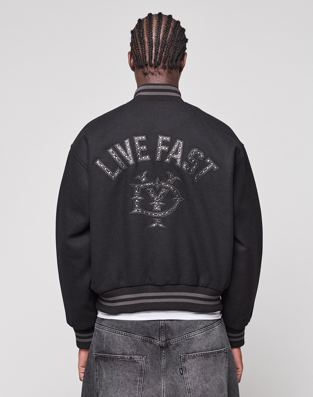 A person with braided hair is seen from behind, wearing the LIVE FAST DIE YOUNG Live Fast College Jacket with a Nieten Patch Logo on the back and metal zipper, paired with regular fit black jeans. | model