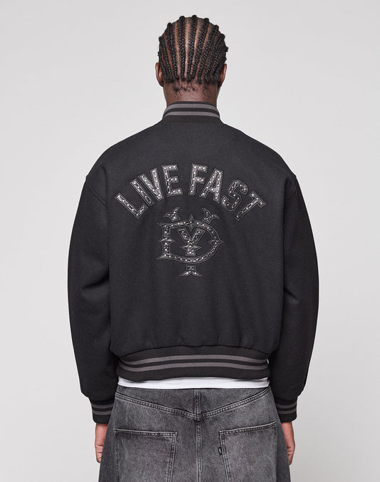 A person with braided hair is seen from behind, wearing the LIVE FAST DIE YOUNG Live Fast College Jacket with a Nieten Patch Logo on the back and metal zipper, paired with regular fit black jeans. | model