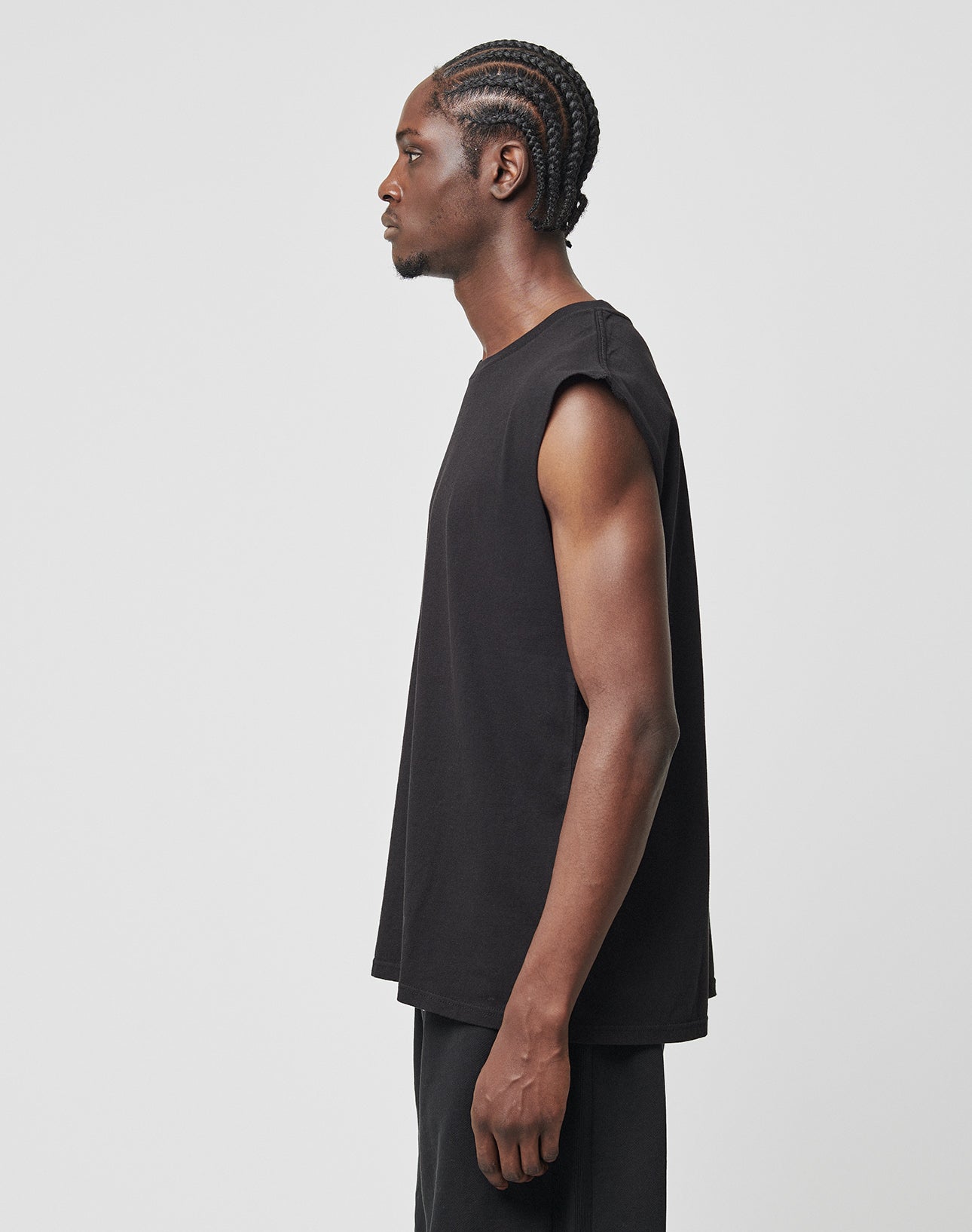 A man with braided hair stands in profile against a plain background, wearing the LIVE FAST DIE YOUNG Basic Muscle Shirt in black—a loose fit, sleeveless top made from 100% Baumwolle with deep armholes for comfort—paired with black pants.