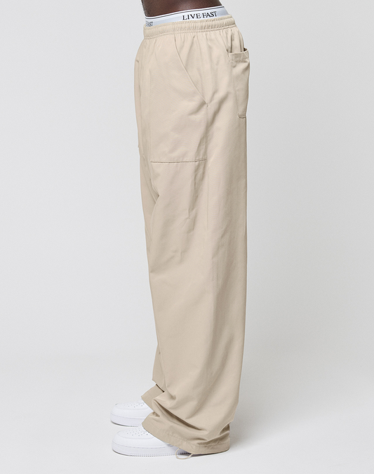 A person in LIVE FAST DIE YOUNG Baggy Nylon Pants with Aufgesetzte Taschen and Elastikbund, white sneakers, and visible white underwear featuring a LIVE FAST waistband, stands against a plain light background.