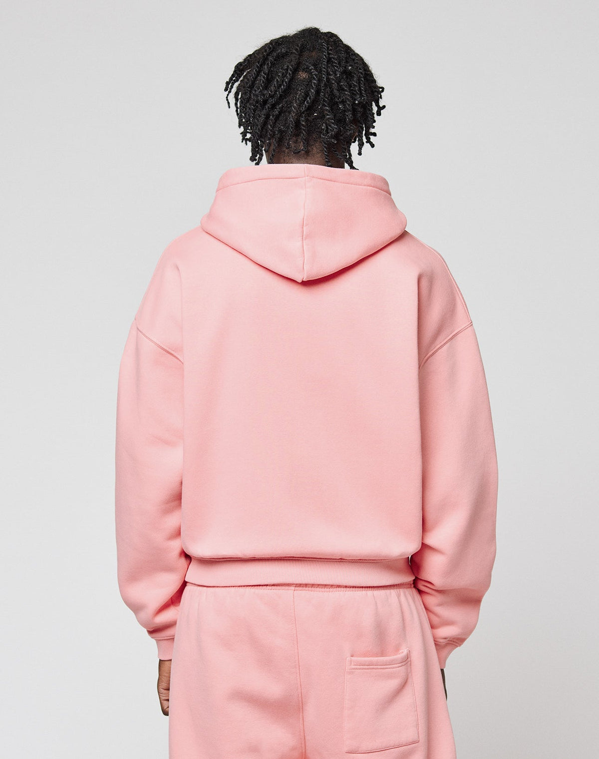 A person with short, twisted hair faces away from the camera, wearing a light pink Basic Hooded Zip and matching sweatpants by LIVE FAST DIE YOUNG, featuring logo branding on the back pocket against a plain light gray background.