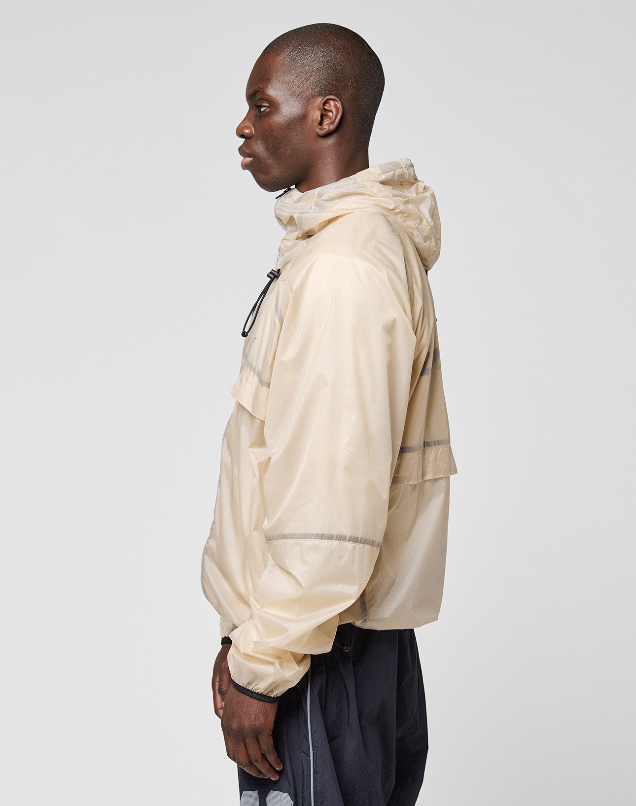 A man in profile wears the LIVE FAST DIE YOUNG Live Fast Nylon Jacket in light beige with reflective print and dark pants, set against a plain light gray background.