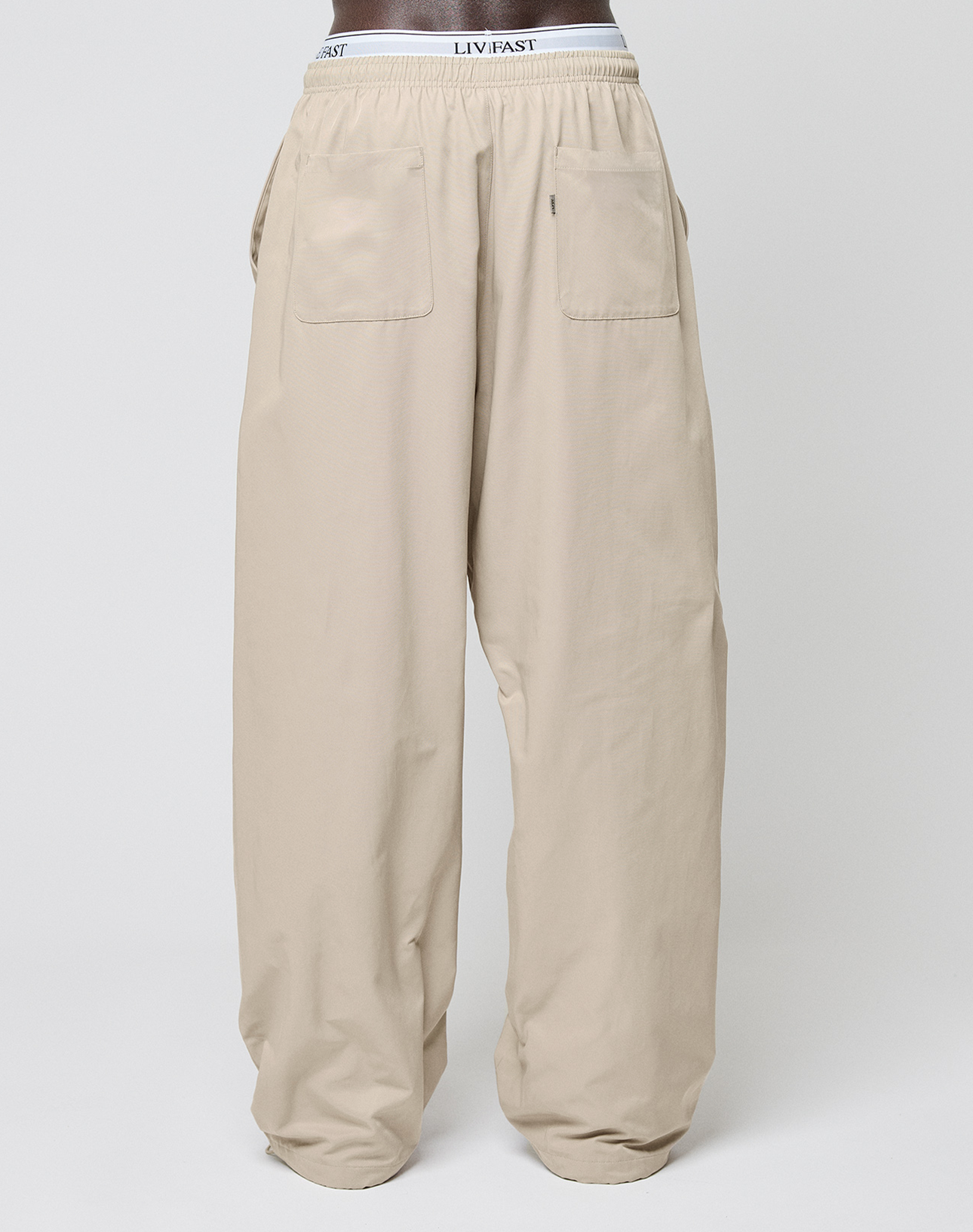Beige Baggy Nylon Pants von LIVE FAST DIE YOUNG im Wide Fit mit zwei aufgesetzten Fronttaschen und elastischem Bund samt sichtbarem LIV FAST Branding, präsentiert an einer Schaufensterpuppe vor schlicht weißem Hintergrund.