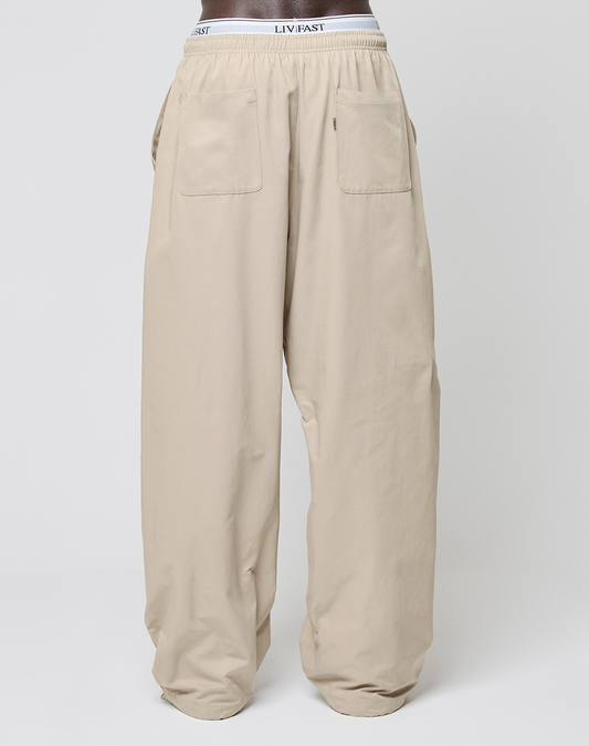 Beige Baggy Nylon Pants von LIVE FAST DIE YOUNG im Wide Fit mit zwei aufgesetzten Fronttaschen und elastischem Bund samt sichtbarem LIV FAST Branding, präsentiert an einer Schaufensterpuppe vor schlicht weißem Hintergrund.