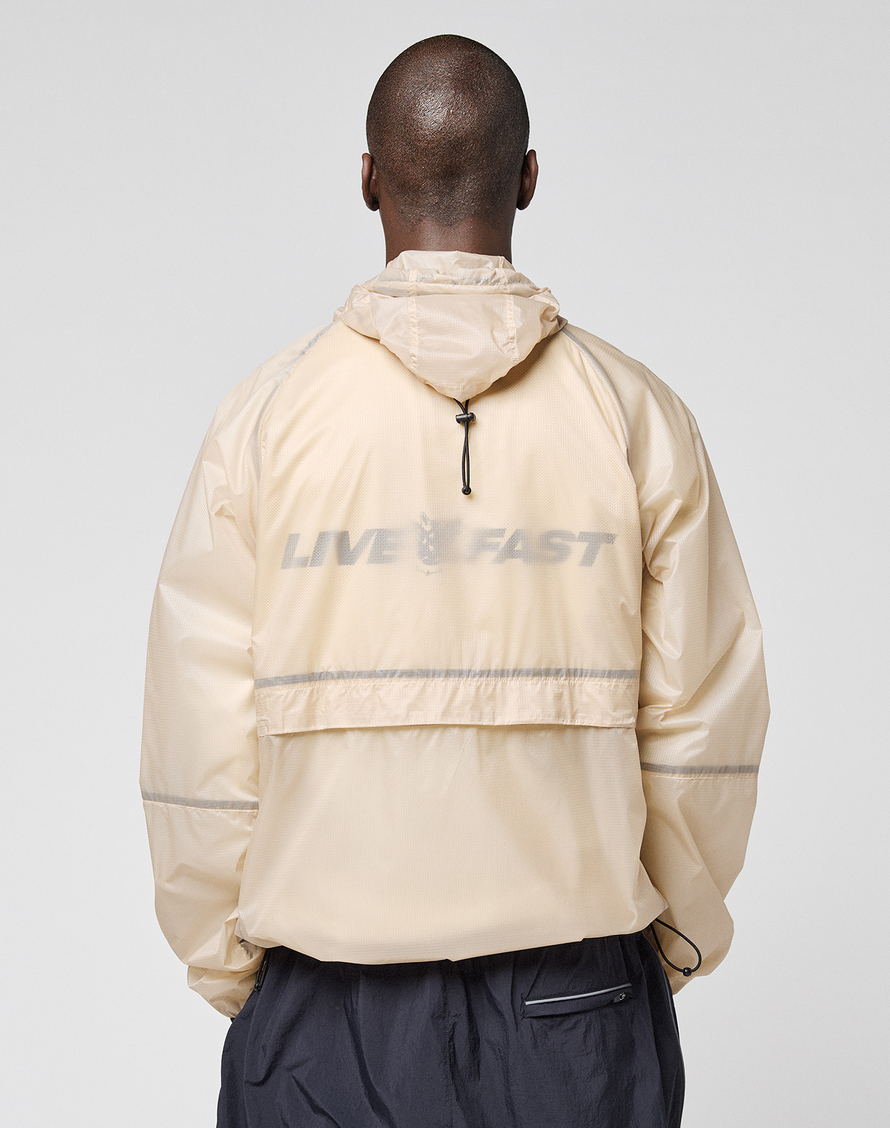 A person is shown from behind wearing the LIVE FAST DIE YOUNG Live Fast Nylon Jacket in light beige, featuring a reflective LIVE FAST print on the back, set against a plain light background.