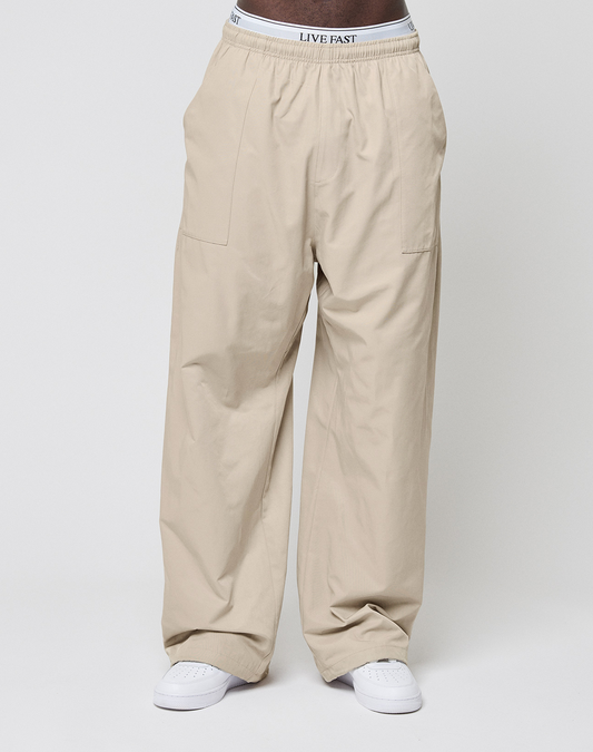 Someone wearing LIVE FAST DIE YOUNG Baggy Nylon Pants in beige with an elastic waistband, paired with white sneakers and visible LIVE FAST branded underwear, stands against a plain light background.