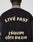 A person wearing the LFDY x Schott NYC Varsity Jacket by LIVE FAST DIE YOUNG, a limited edition black varsity with cream chenille patches and LIVÉ FAST & LÉQUIPE CÔTE DAZUR text on the back, head turned to show only jacket and neck.