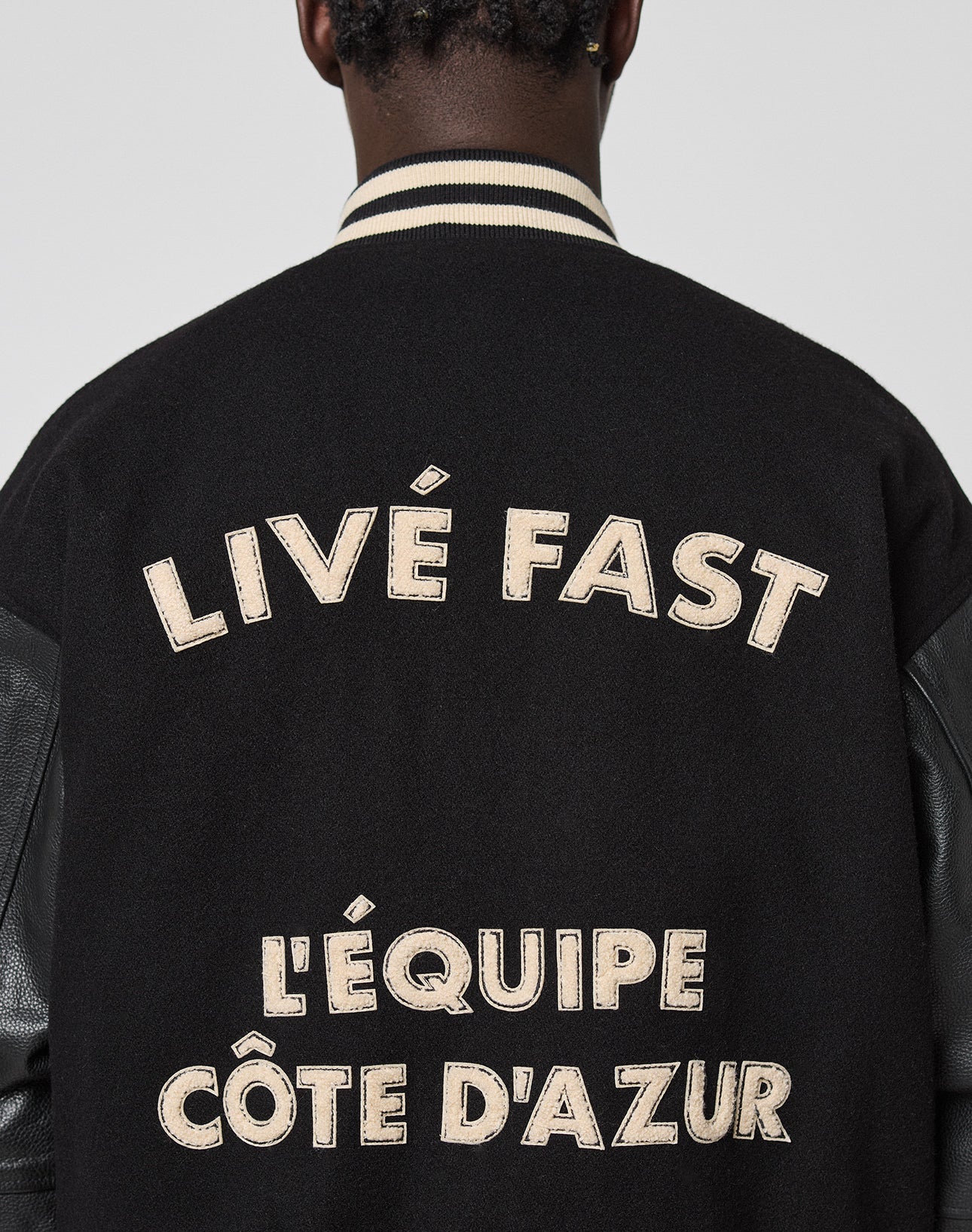 A person wearing the LFDY x Schott NYC Varsity Jacket by LIVE FAST DIE YOUNG, a limited edition black varsity with cream chenille patches and LIVÉ FAST & LÉQUIPE CÔTE DAZUR text on the back, head turned to show only jacket and neck.
