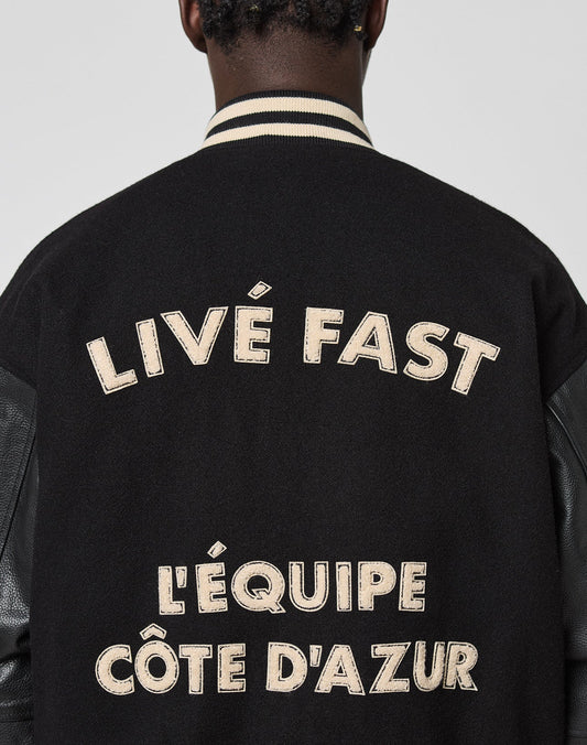 A person wearing the LFDY x Schott NYC Varsity Jacket by LIVE FAST DIE YOUNG, a limited edition black varsity with cream chenille patches and LIVÉ FAST & LÉQUIPE CÔTE DAZUR text on the back, head turned to show only jacket and neck.
