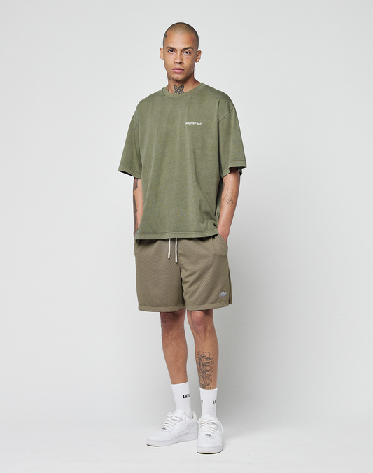 A person stands against a plain white background wearing an olive green oversized t-shirt, LIVE FAST DIE YOUNG Basic Mesh Shorts in olive with an elastic waistband and subtle logo patch, white crew socks, and white sneakers for a casual look.