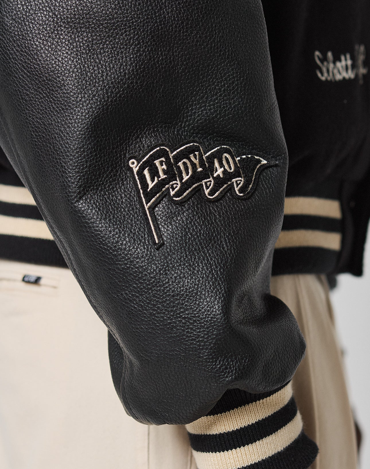 Close-up of the LFDY x Schott NYC Varsity Jacket by LIVE FAST DIE YOUNG, featuring a black leather sleeve with a waving flag patch reading “LF DY 40” and black and beige striped cuffs; beige pants are partially visible.