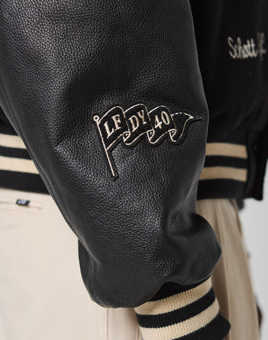 Close-up of the LFDY x Schott NYC Varsity Jacket by LIVE FAST DIE YOUNG, featuring a black leather sleeve with a waving flag patch reading “LF DY 40” and black and beige striped cuffs; beige pants are partially visible.
