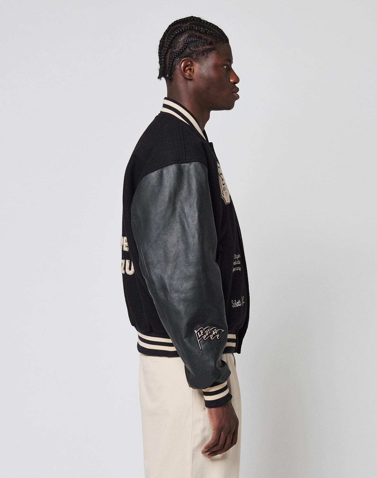 A person with braided hair stands in profile, wearing the LFDY x Schott NYC Varsity Jacket by LIVE FAST DIE YOUNG and cream pants against a plain light background.