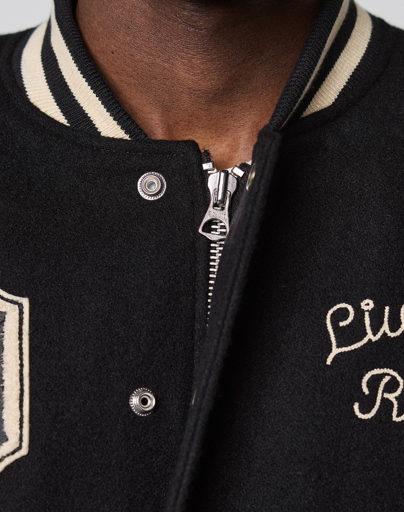 Close-up of a person in the LFDY x Schott NYC Varsity Jacket by LIVE FAST DIE YOUNG, featuring a black design with a white striped collar and embroidered text. Only the lower face and upper torso are shown; the jacket is partially unzipped.