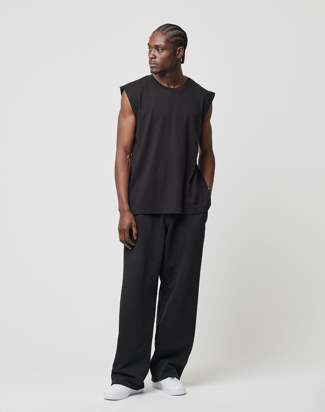 A man with braided hair wears the LIVE FAST DIE YOUNG Basic Muscle Shirt, loose black pants, and white sneakers. He stands against a plain white background, looking left with one hand in his pocket.